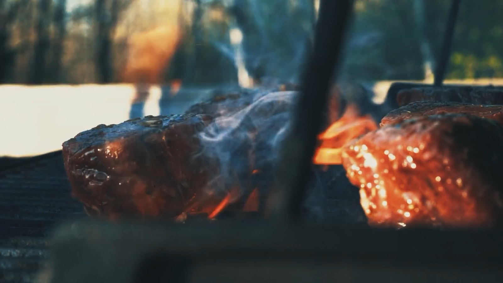 Fleisch auf dem Grillrost über glühender Kohle, aufsteigender Rauch, Abendsonne über einem See im Hintergrund
